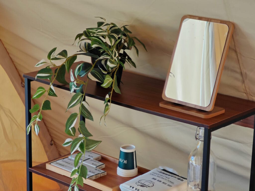 Stylish shelving with glamping essentials including a potted plant, mirror, reusable water bottle, portable fan, kettle, neatly arranged shower cap and towel, representing must-have items for a luxury glamping trip at IKAN Glampsite in Janda Baik.