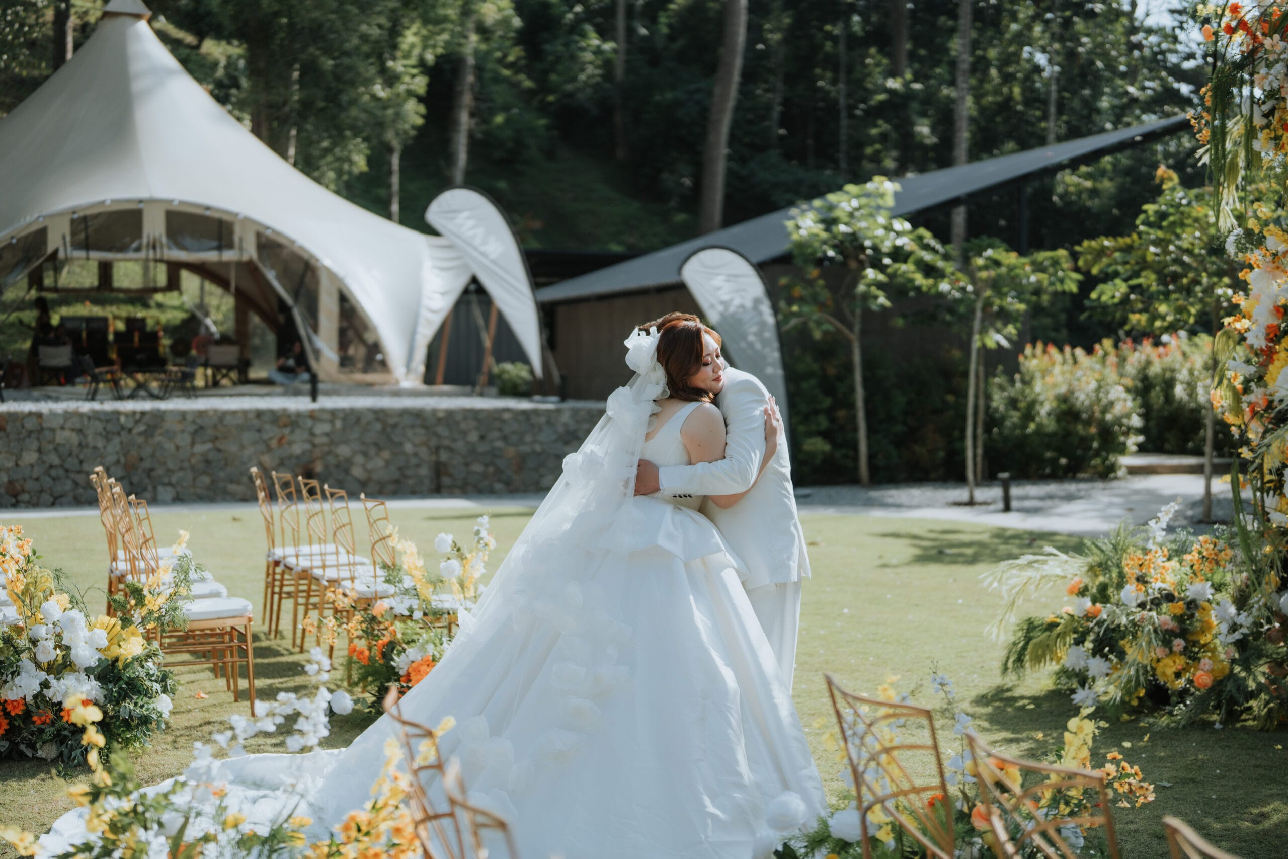Bride and groom embrace at outdoor glamping wedding venue in Janda Baik, Bentong, surrounded by white tents, floral arrangements, and lush forest backdrop.