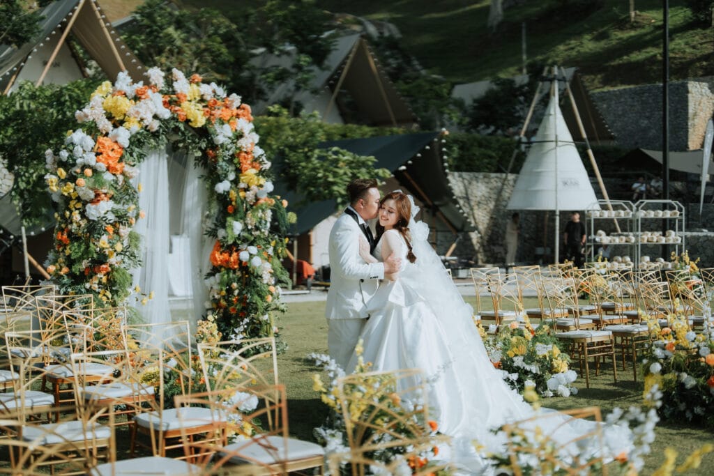 Bride and groom posing under floral wedding arch in elegant outdoor garden ceremony surrounded by gold chairs and vibrant flowers