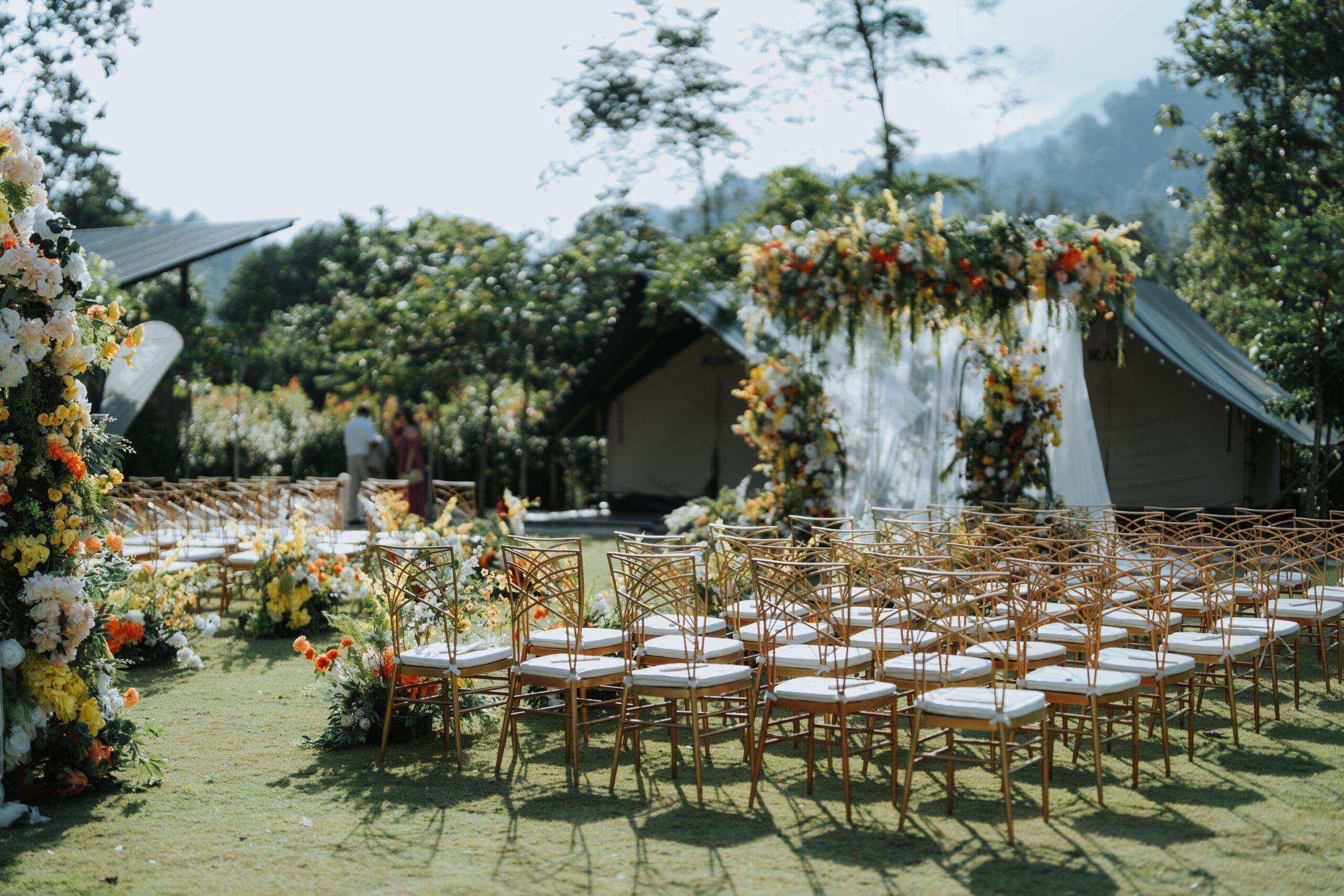 A beautifully decorated outdoor wedding setup at IKAN Glampsite featuring gold chairs, vibrant floral arrangements, and a garden aisle surrounded by lush greenery.