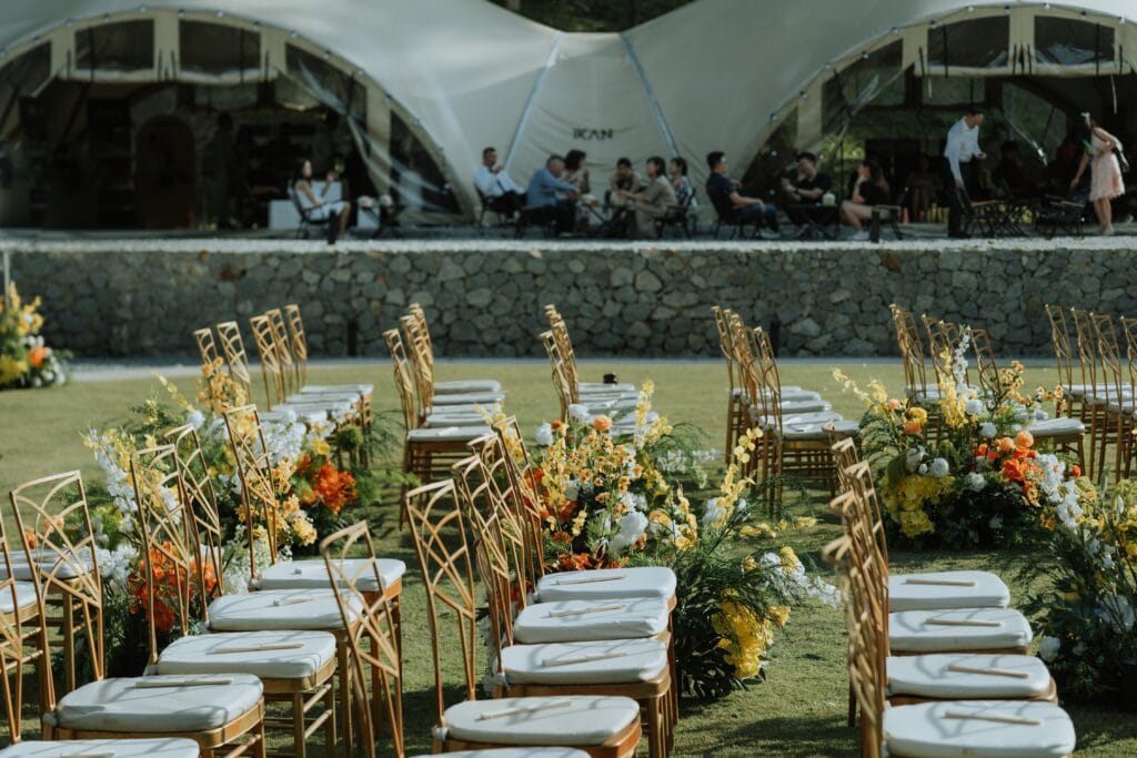 Garden wedding ceremony seating arranged on a lawn with colourful fresh flowers, with IKAN Glampsite’s signature tent structures visible in the background.