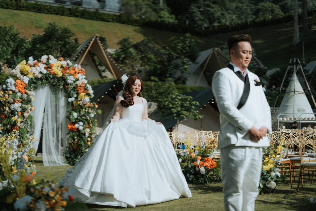 A bride walking past a vibrant floral arch during her outdoor wedding ceremony at IKAN Glampsite, surrounded by greenery and golden-hour lighting.